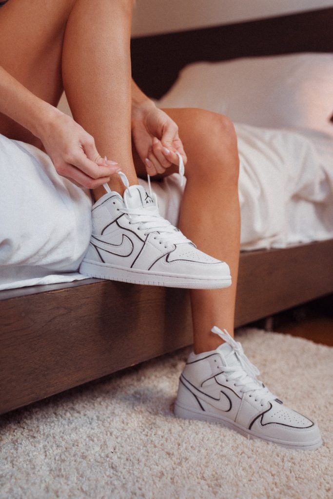A photo of a person's legs hanging off the side of a bed, as they tie the laces on one of their white sports shoes.