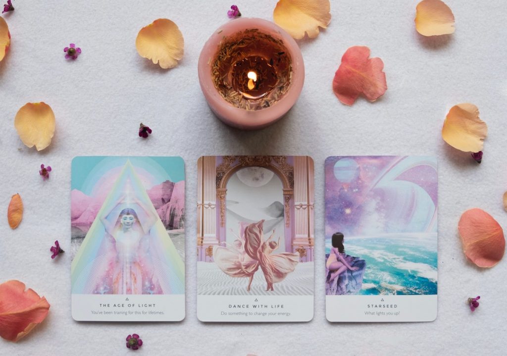 A photo of three oracle cards spread out amidst scattered flower petals, with a candle burning in the top middle.