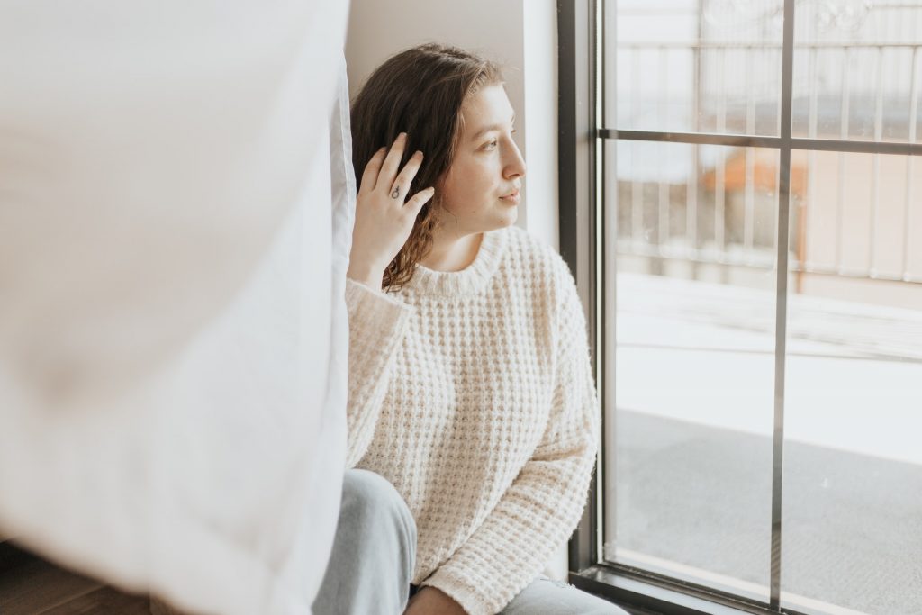 A woman sits near a window, looking outside. She's wearing a comfortable cream colored knit sweater. Dressing comfortably is one of my top period tips.
