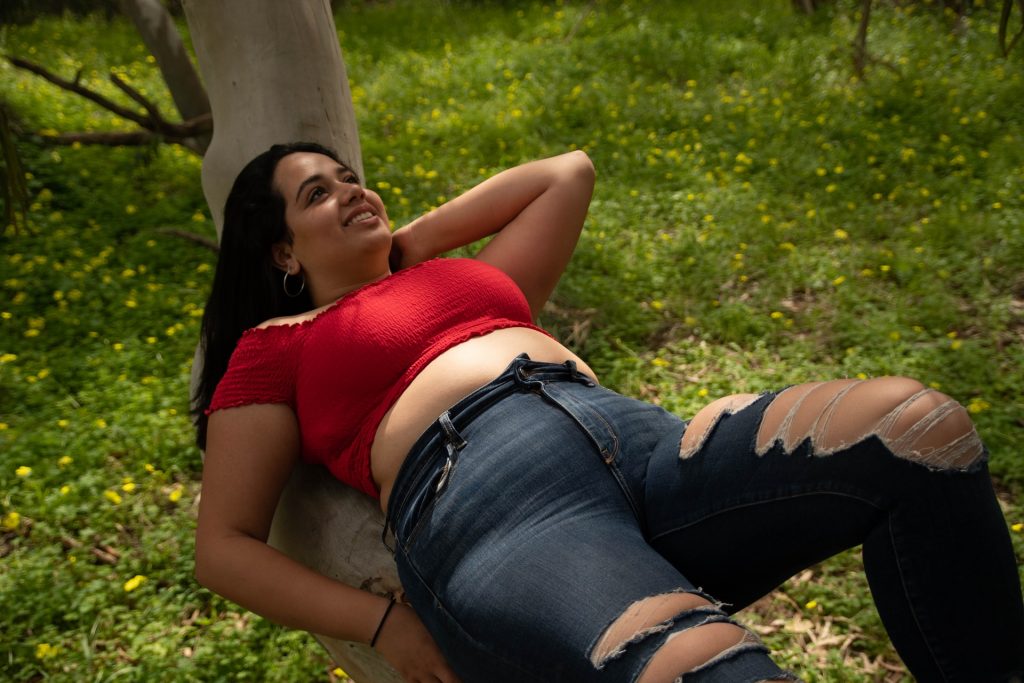 A person in a red shirt and blue jeans, leaning back on a tree branch, looking up with a smile.