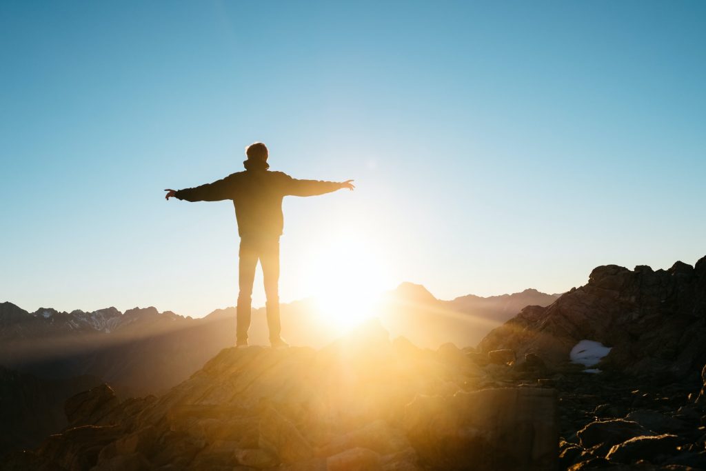 A photo of a person standing on a mountain with their arms spread wide, looking out at the bright sun. I am statements can be empowering.