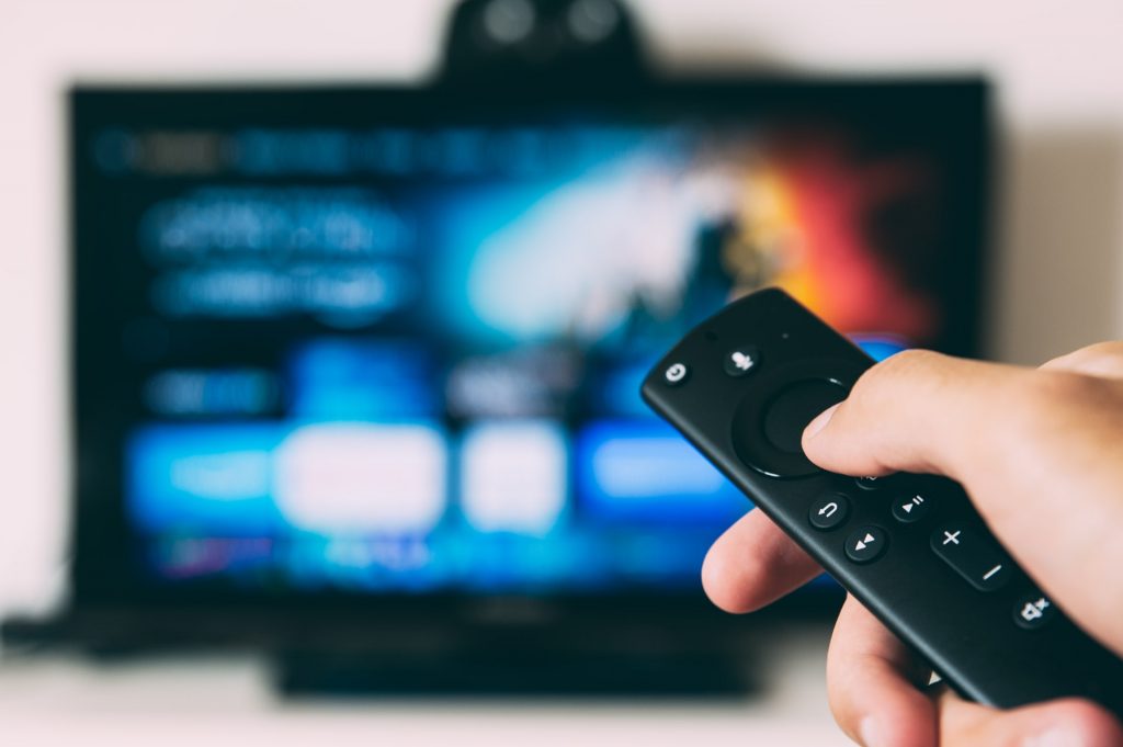 A photo of a TV remote in someone's hand, with a blurred TV screen in the background. You can travel from home by watching films and TV from other parts of the world.