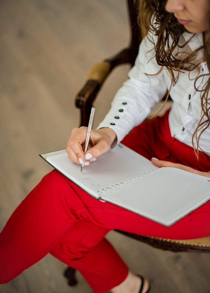 A person in a white shirt and red pants sits in a chair and writes in a journal.