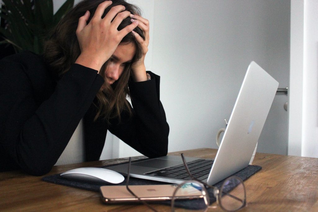 A person sits at a table with their laptop, holding their head in their hands and looking overwhelmed. Workplace perfectionism can be battled with affirmations for perfectionism.
