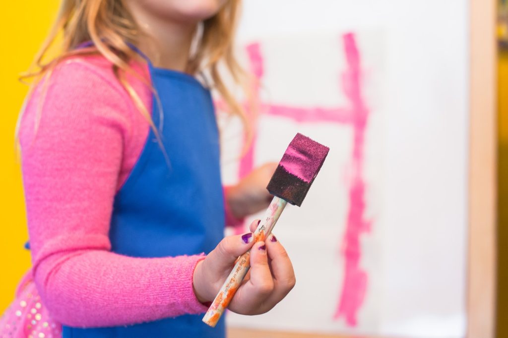 A child in a pink shirt and blue apron holds a painting sponge with pink paint on it.