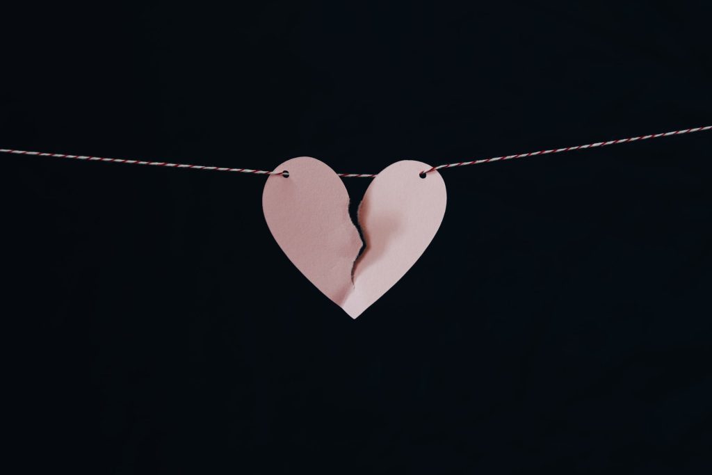 A photo of a pink paper heart on a string that is torn in half, amidst a black background.