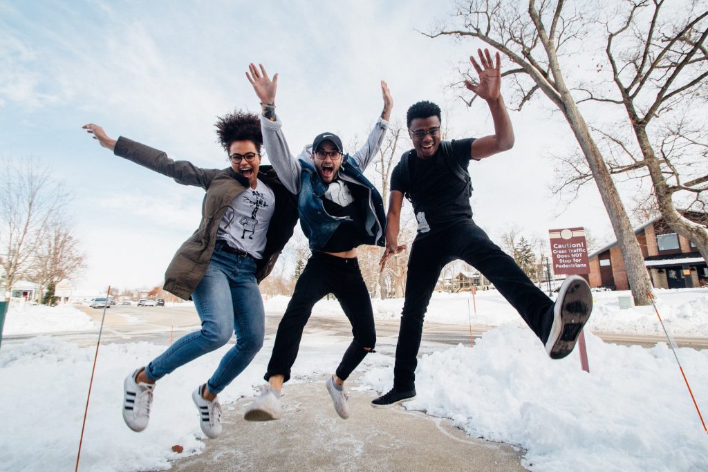 A photo of three friends, outside on a snowy street, caught mid-air as they jump with big smiles on their faces.