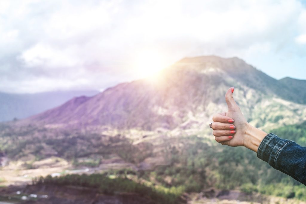 A photo of a beautiful mountain with the sun shining bright behind it, while a person to the right side holds their hand out in a thumbs up position.