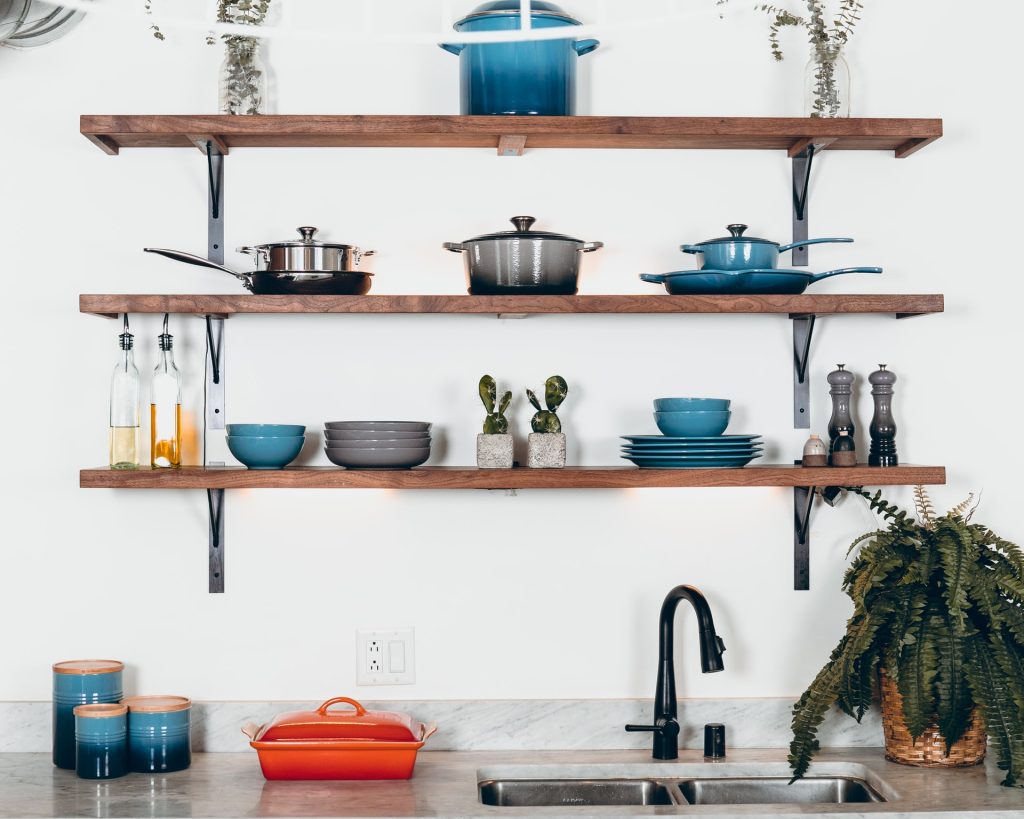A photo of a kitchen sink with wooden open shelving on the wall above it, with pots and pans and dishes organized neatly. 
