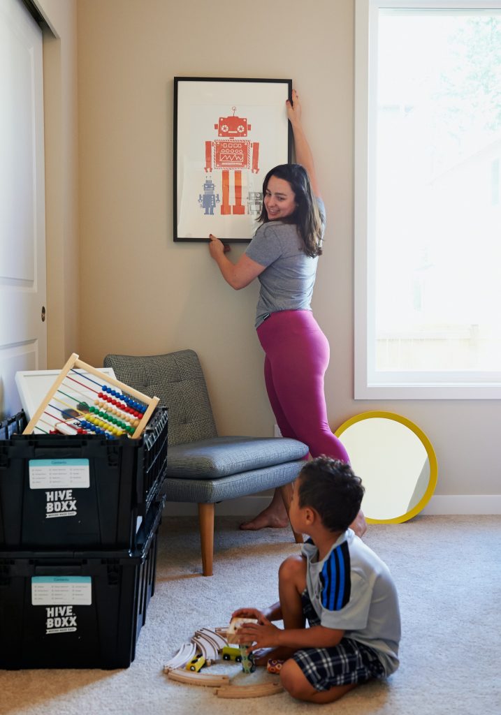 A photo of an adult smiling and hanging up a picture, as a child sits on the floor near a stack of moving boxes.
