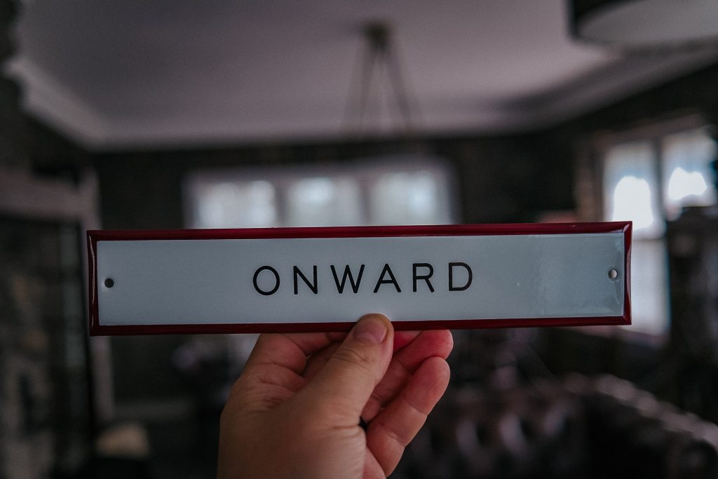A photo of a hand holding up a small sign that says "Onward". Now that you know how to organize your life in one week, the key is to stick with it!