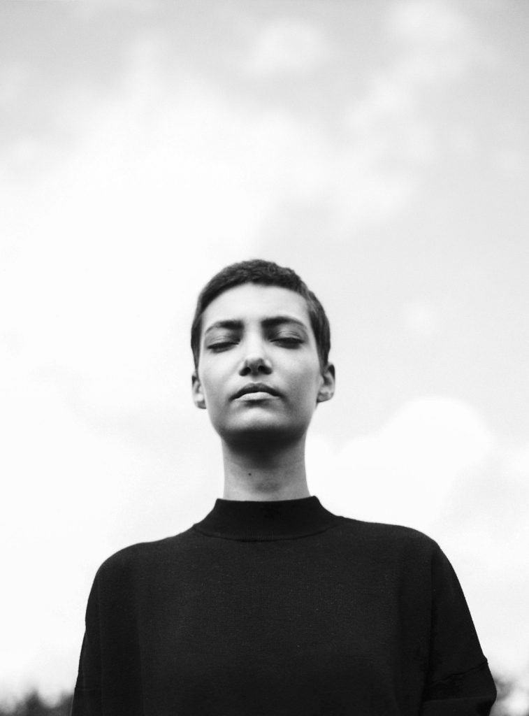 A black and white photo of a person with short hair standing amidst a cloudy sky with their eyes closed, a peaceful expression on their face.