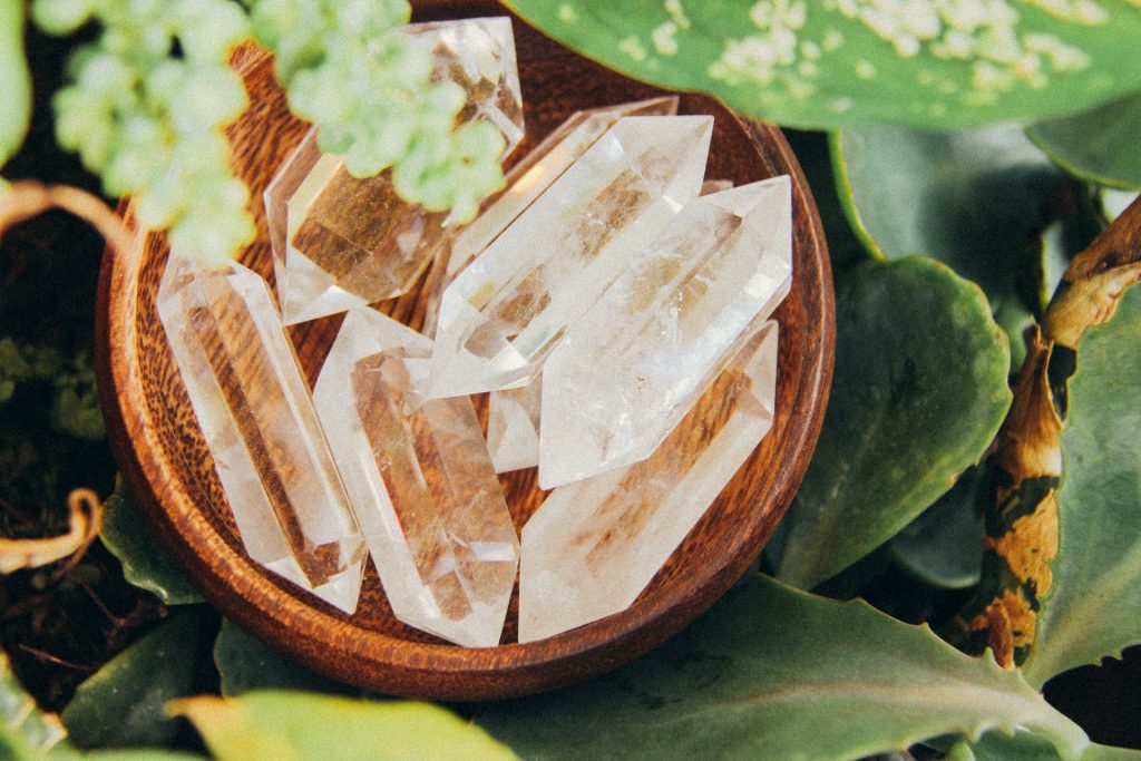 A photo of a wooden bowl full of clear quartz crystals, amidst a background of green leaves. Categories of affirmations for clear quartz include clarity and manifestation.