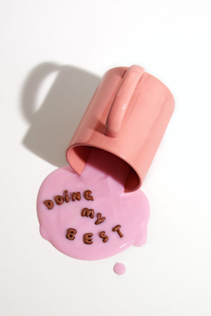 A photo of a coral pink mug tipped on its side against a white background, pink contents spilling out of it and brown alphabet cereal pieces arranged to spell the words "Doing my best".