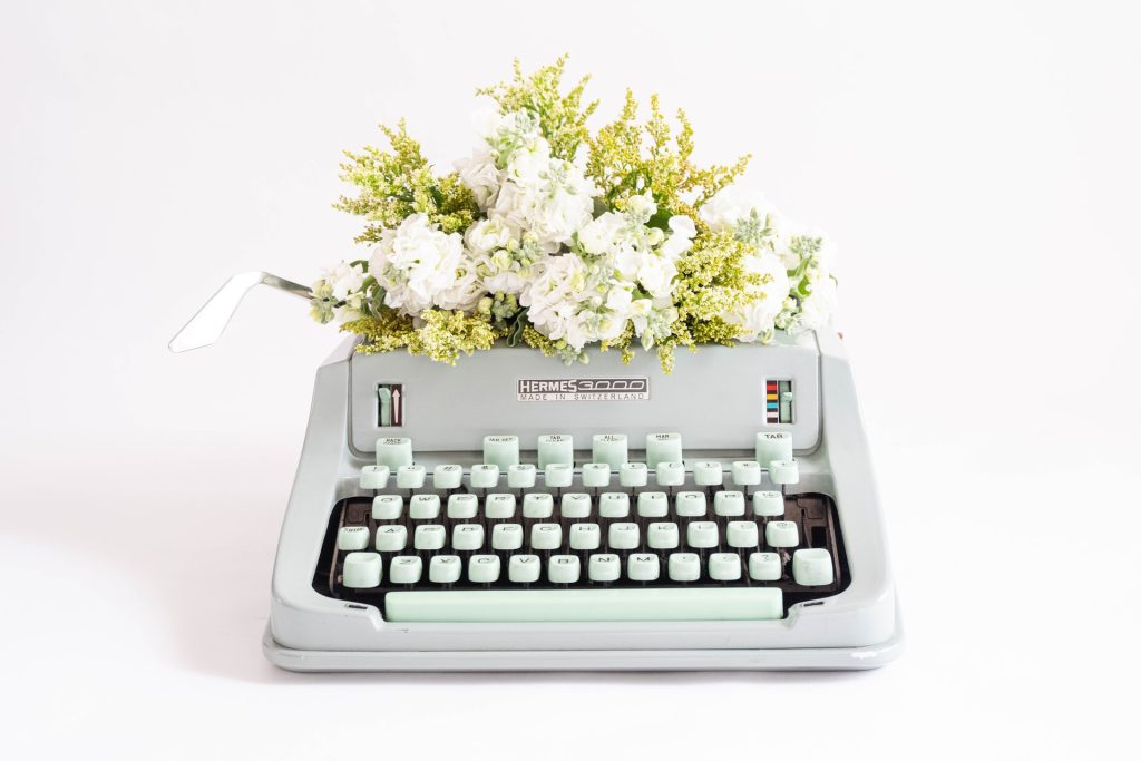 A photo of a grey typewriter with mint green keys, sitting amidst a white background. There are white and yellow green flowers coming out of the paper holder in the typewriter. Positive affirmations for creativity must include some for writers!