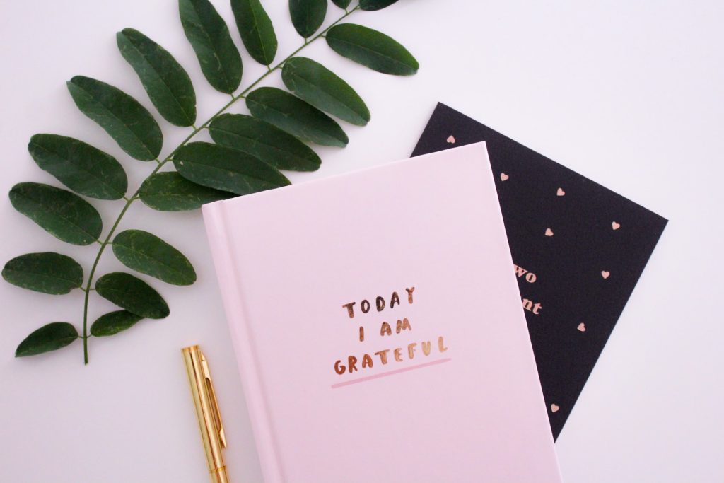 A photo of a pink journal that says Today I Am Grateful on the cover. Beside it rests a gold pen, and on the other side, covered mostly by the journal, is some sort of black card or art print with pink hearts on it. There is a green twig of leaves in the upper left corner of the photo.