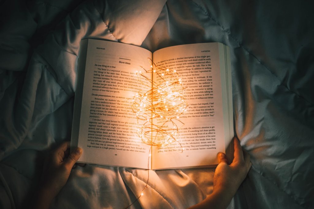 A photo of a person holding a book open on their bed, with a string of bright fairy lights bunched up between the pages, illuminating the words with a soft orange glow.