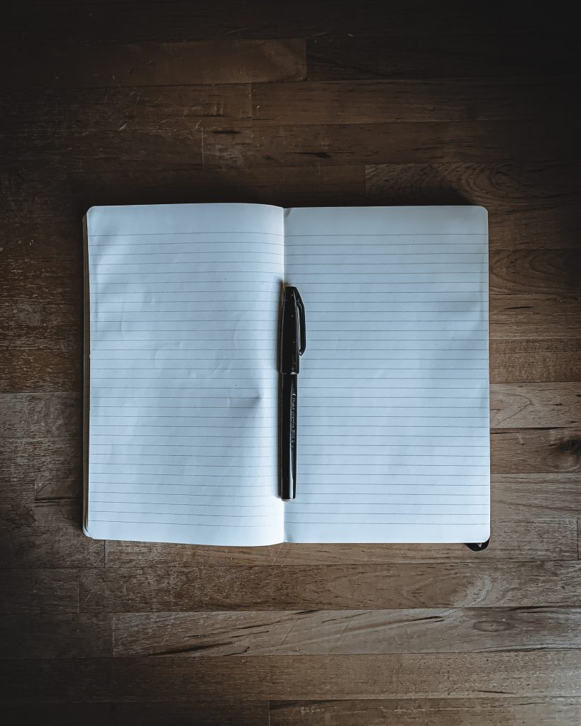A photo of a blank notebook spread open on a wooden table, a black pen lying in the center of it. 