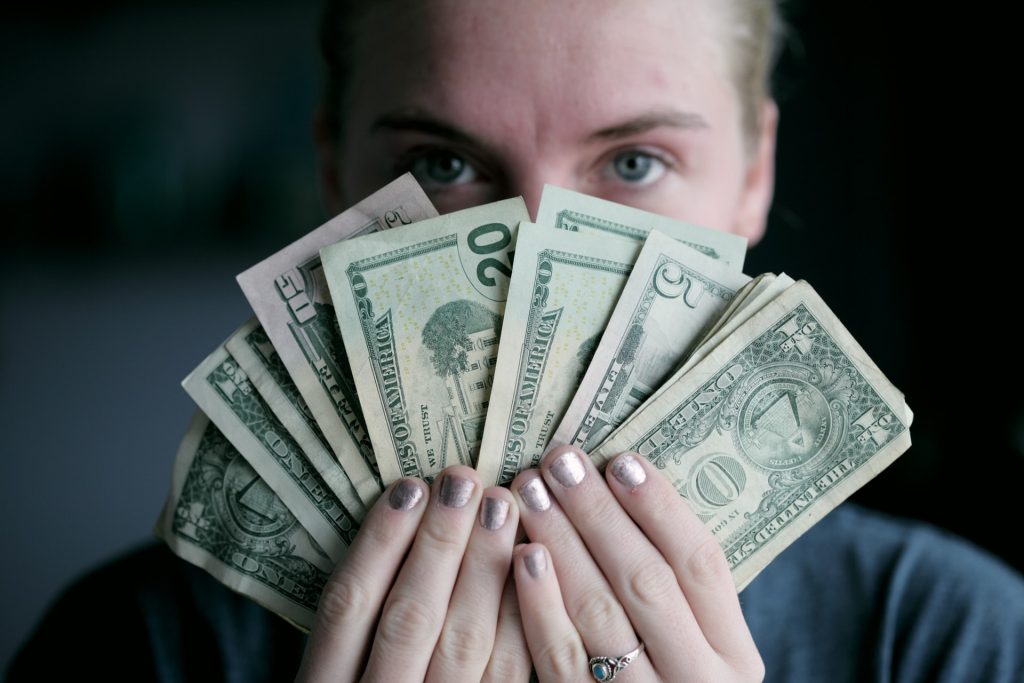 A photo of a person holding paper money in front of their face, their eyes peeking out from behind the fan of one, five, twenty, and fifty dollar bills.